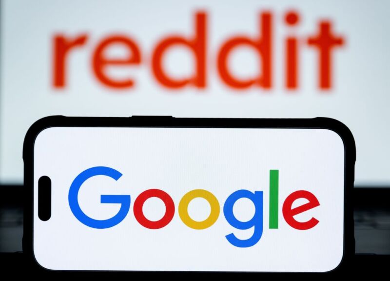 Image 1: Google is displayed on a mobile phone screen in front of logo of Reddit is displayed on a computer screen i